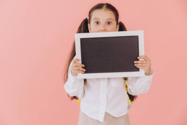 Elinde karatahta tutan komik küçük kız yüzünün yarısını kaplamış ve altından dışarı bakıyor, pembe arka planda kopya alanı var.