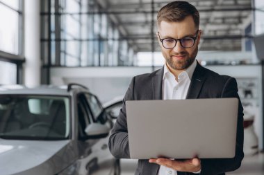 Araba galerisinde dizüstü bilgisayar kullanan gözlüklü ve takım elbiseli modern sakallı bir adam.