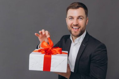 Sakallı mutlu gülen adam elinde bir hediye tutuyor ve onu gri bir arkaplanda açıyor. Sevgililer günü, Noel ve yeni yıl konsepti.