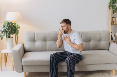 Hasta adam oturma odasında öksürürken göğsünü ağrı içinde tutuyor. Boğaz ağrısı ve öksürük, akciğer ağrısı olan bir adam, sağlık sorunları konsepti.