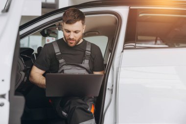 Otomotiv teknisyeni bilgisayarda tanılama yazılımı çalıştırıyor. Tamirci, oto servis garajında aracın tanılama testini yaparken arabada oturup dizüstü bilgisayarla çalışıyor..
