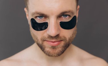 Yakışıklı sakallı adamın yakın plan portresi. Gözlerinin altına siyah bantlar yapıştırıyor. Gri arka plandaki görünüşünü önemseyen çekici adam.