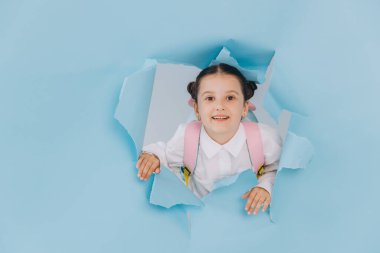 Küçük tatlı mutlu okul kızı mavi kağıttaki yırtık delikten her yere bakıyor.