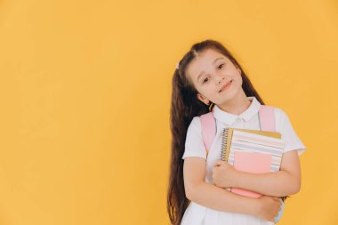 Sırt çantalı, sarı arka planda defterleri ve kitapları olan beyaz okul üniformalı küçük tatlı kız.