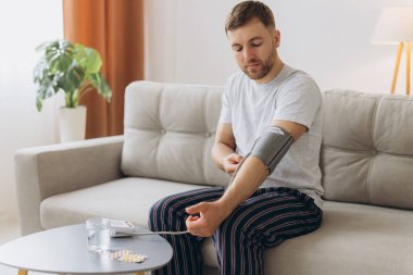 Evdeki kanepede oturan genç beyaz adam, kan basıncını ölçüyor. Modern tonometre kullanan sakallı adam stres ve baş ağrısından kaynaklanan hipertansiyon yaşıyor.