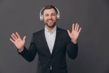 Mutlu, sakallı, takım elbiseli bir işadamı gri bir stüdyonun arka planında dans ediyor ve kulaklıkları dikkatle dinliyor.