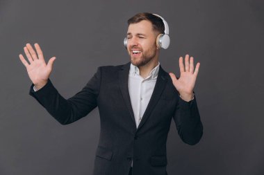 Mutlu, sakallı, takım elbiseli bir işadamı gri bir stüdyonun arka planında dans ediyor ve kulaklıkları dikkatle dinliyor.