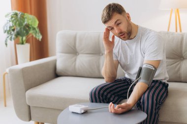 Evdeki kanepede oturan genç beyaz adam, kan basıncını ölçüyor. Modern tonometre kullanan sakallı adam stres ve baş ağrısından kaynaklanan hipertansiyon yaşıyor.