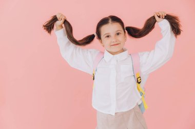 Sırt çantasıyla pembe arka planda uzun saçlarını tutan okul üniformalı sevimli, küçük bir kız.