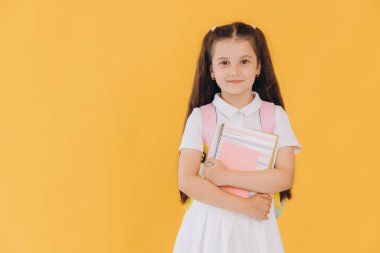 Sırt çantalı, sarı arka planda defterleri ve kitapları olan beyaz okul üniformalı küçük tatlı kız.