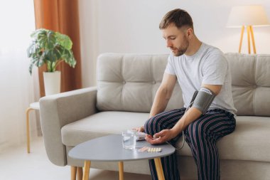 Evdeki kanepede oturan genç beyaz adam, kan basıncını ölçüyor. Modern tonometre kullanan sakallı adam stres ve baş ağrısından kaynaklanan hipertansiyon yaşıyor.