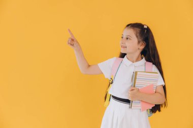 Sırt çantasında defterler olan ve sarı arka planda bir şey gösteren beyaz okul üniformalı tatlı birinci sınıf öğrencisi kız.