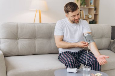 Evdeki kanepede oturan genç beyaz adam, kan basıncını ölçüyor. Modern tonometre kullanan sakallı adam stres ve baş ağrısından kaynaklanan hipertansiyon yaşıyor.