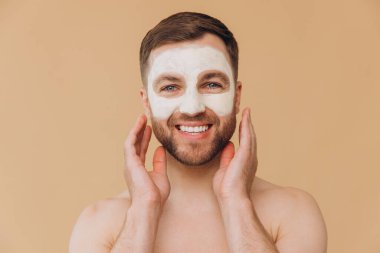 Tıraşsız, gülümseyen bir adamın güzel portresi bej renkli stüdyo arka planına yüz bakımı maskesi takıyor. Dermatoloji, lüks spa prosedürleri. Erkek güzellik anlayışı, kozmetik ürünler..