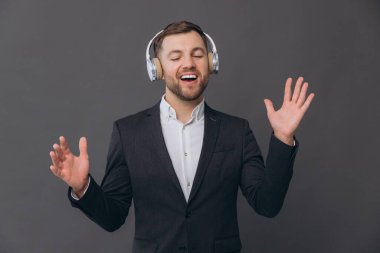 Mutlu, sakallı, takım elbiseli bir işadamı gri bir stüdyonun arka planında dans ediyor ve kulaklıkları dikkatle dinliyor.