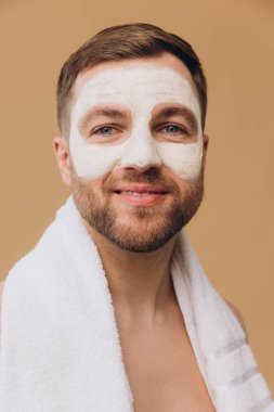 Bej stüdyo arka planına yüz maskesi takan, boynu havluyla gezen, traşsız, gülümseyen bir adamın güzel portresi. Dermatolojik sağlık, lüks kaplıca tedavileri. Erkeklerin güzellik anlayışı, kişisel bakım kozmetik.