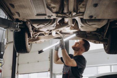 Yoğunlaştırılmış araba servisi çalışanının otomobil kontrol ekipmanlarına bakın. Üniformalı ve eldivenli erkek tamirci arabanın altında duruyor, lambayı tutuyor ve kırılma noktası arıyor.