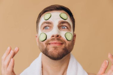 Tıraşsız, komik, boynunda havluyla gezen, bej renkli stüdyo arka planına salatalık şeritleri takan mutlu bir adamın güzel portresi. Dermatolojik sağlık, lüks kaplıca tedavileri.