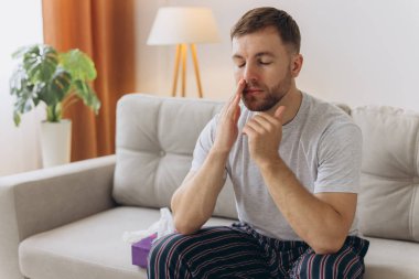 Sakallı, hasta, üzgün bir adam evdeki kanepede oturuyor. Burun nezlesi salgını, koronavirüs salgını, çoban salgını, hapşırma....