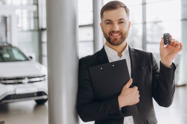 Gülümseyen araba satıcısı araba galerisinde duruyor ve satılmaya hazır yeni bir arabanın anahtarlarını gösteriyor.