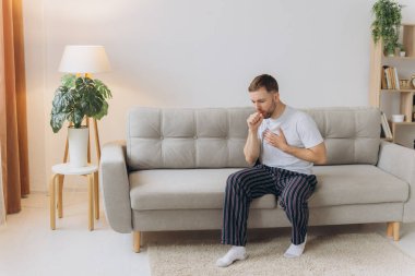 Hasta adam oturma odasında öksürürken göğsünü ağrı içinde tutuyor. Boğaz ağrısı ve öksürük, akciğer ağrısı olan bir adam, sağlık sorunları konsepti.