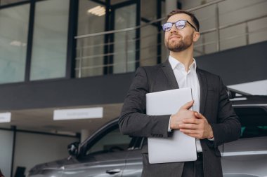 Araba galerisinde dizüstü bilgisayar kullanan gözlüklü ve takım elbiseli modern sakallı bir adam.