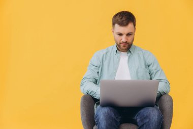 Tıraşsız, naneli gömlekli bir adam koltukta oturur, beklemede kalır, bilgisayardaki işi kullanır, sarı arka plandaki stüdyoda izole edilir..