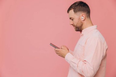 Yan görünüm mutlu beyaz sakallı adam pembe gömlek giyer günlük kıyafetler giyer kulaklıkla müzik dinler basit pastel açık pembe arka plan stüdyo portresi üzerinde izole edilmiş telefona bakar