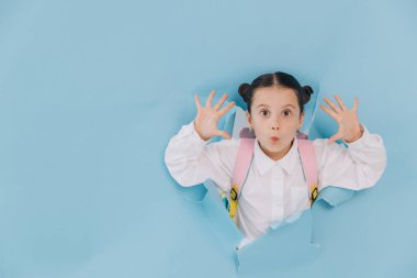 Küçük tatlı mutlu liseli kız yırtık deliğe bakıyor ve mavi kağıtta komik bir surat yapıyor.