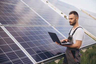 Makinist ya da inşaat işçisi sakallı adam güneş paneli durumunu kontrol etmek için dizüstü bilgisayar kullanıyor. Hücre santrali. Gelişmiş gerçeklik kavramı. Gelecekçi teknoloji