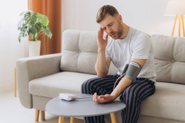 Evdeki kanepede oturan genç beyaz adam, kan basıncını ölçüyor. Modern tonometre kullanan sakallı adam stres ve baş ağrısından kaynaklanan hipertansiyon yaşıyor.