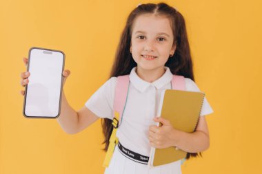 Okula geri döndüm: okul kızı cep telefonunu boş ekranla gösteriyor ve sarı arka planda defteri tutuyor, teknoloji kavramını gözler önüne seriyor.