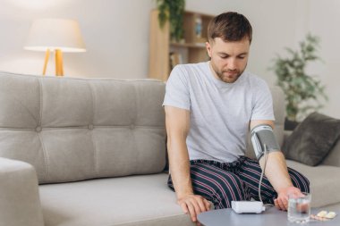 Evdeki kanepede oturan genç beyaz adam, kan basıncını ölçüyor. Modern tonometre kullanan sakallı adam stres ve baş ağrısından kaynaklanan hipertansiyon yaşıyor.