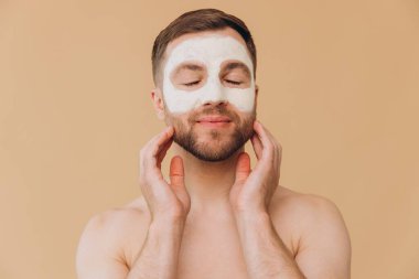 Tıraşsız, gülümseyen bir adamın güzel portresi bej renkli stüdyo arka planına yüz bakımı maskesi takıyor. Dermatoloji, lüks spa prosedürleri. Erkek güzellik anlayışı, kozmetik ürünler..