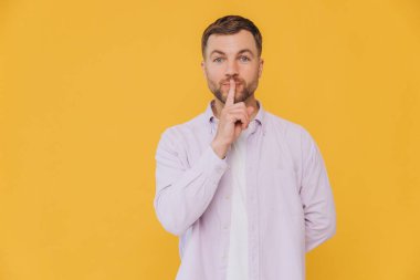 Mor gömlekli, ağzının yanında parmağını tutan ve sarı arka planda izole edilmiş sevimli, tıraşsız bir adam.