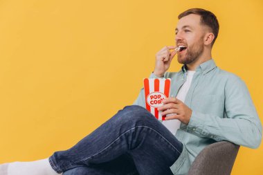 Nane gömlekli mutlu 30 'lu yaşlarda bir sandalyede oturup patlamış mısır kovanlı film ya da sarı arka plan stüdyo portresi üzerinde izole edilmiş futbol maçı izleriz. Halk yaşam tarzı konsepti