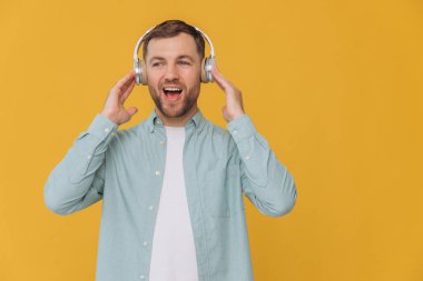 Tıraşsız, temiz gömlekli, kulaklıkla müzik dinleyen ve sarı arka plandaki fotokopi alanını gösteren yakışıklı bir adam.