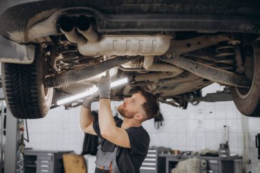 Yoğunlaştırılmış araba servisi çalışanının otomobil kontrol ekipmanlarına bakın. Üniformalı ve eldivenli erkek tamirci arabanın altında duruyor, lambayı tutuyor ve kırılma noktası arıyor.