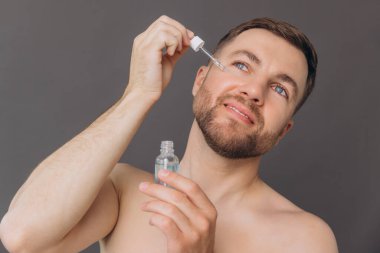 Yakışıklı, gülümseyen sakallı bir adam gri bir stüdyoda güzellik ve cilt rutini sırasında yanağında serum olan pipeti tutuyor..