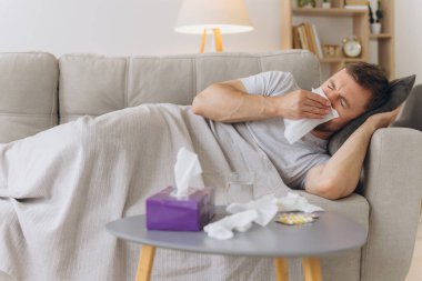 Evdeki kanepede sıcak bir battaniyenin altında yatıp kağıt mendille hapşıran hasta, beyaz bir adam. Soğuk algınlığı, grip, mevsimsel kış enfeksiyonları