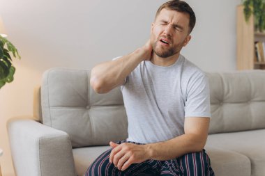 Boyun ağrısı. Kafkasyalı bir adam evdeki oturma odasında boynuna masaj yapıyor. Kemik eklem iltihabı, Sağlık Sorunu Kavramı.