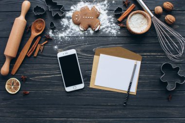 A cozy setup for Christmas baking with gingerbread cookies, cinnamon, and a whisk on a rustic wooden table. Includes a smartphone, rolling pin, and blank note for a festive touch. clipart