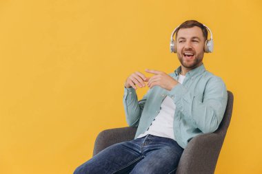 Naneli gömlekli, traşsız yakışıklı bir adam sandalyede oturmuş, kulaklıkla müzik dinliyor sarı arka planda gitar çalıyormuş gibi yapıyor.