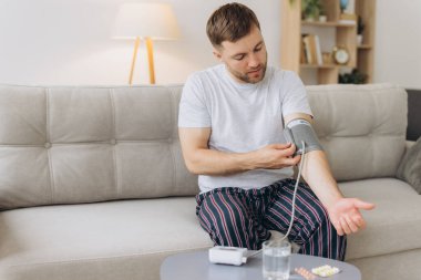 Evdeki kanepede oturan genç beyaz adam, kan basıncını ölçüyor. Modern tonometre kullanan sakallı adam stres ve baş ağrısından kaynaklanan hipertansiyon yaşıyor.