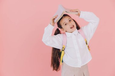 Sırt çantası açık, pembe arka planda kitabı tutan okul üniformalı sevimli, küçük bir kız.