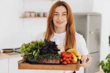 Mutlu, olgun, gülümseyen ve elinde sağlıklı yiyeceklerle evde duran bir kadın.