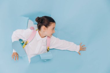 Küçük tatlı, mutlu okul kızı yırtık deliğe bakıyor. Mavi kağıtta kopya alanı.