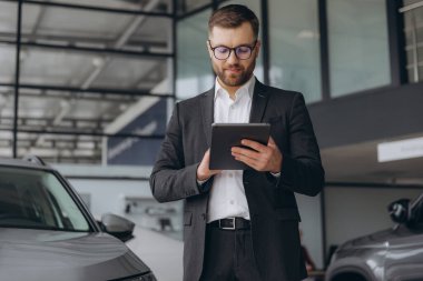 Sakallı, gözlüklü, takım elbiseli, araba tamirhanesinde duran ve elinde tablet tutan dost canlısı araba satıcısı.