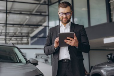 Bearded friendly car seller in glasses and suit standing in car salon and holding tablet clipart