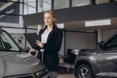 Yakışıklı araba galerisi kadın çalışanı elinde bir klasör tutuyor ve arabanın yanında dururken gülümsüyor.